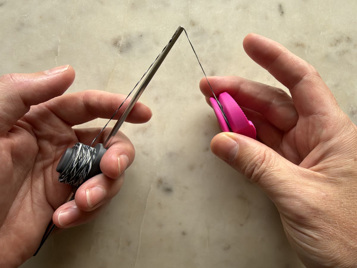 Two hands: the Flosser Wand held in one, the Flosser Ring with floss wound on it in the other, and the floss running between them